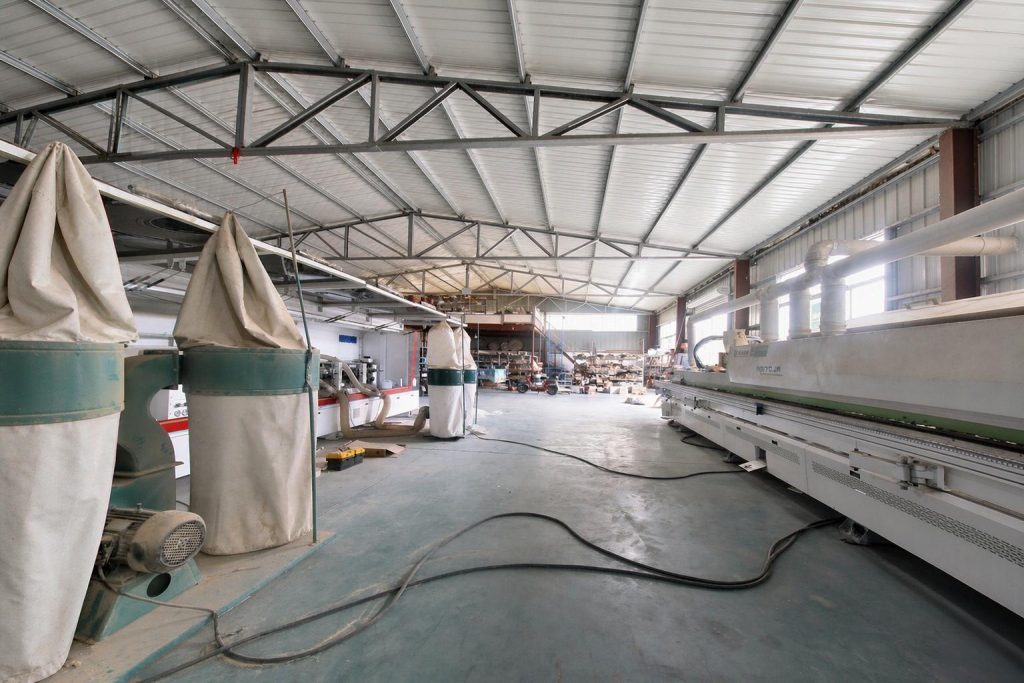 Woodworking workshop with edge banding machinery and dust extraction units under a steel truss factory roof